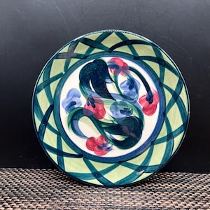 May include: A round ceramic plate with a hand-painted floral design. The plate features a white center with blue, red, and green floral motifs. The rim has a green lattice pattern with blue accents. The plate is on a dark surface.