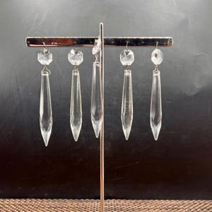 May include: Five clear glass chandelier crystals hanging from a silver-toned metal display stand. Each crystal features a faceted octagonal top piece connected to a long, pointed, tapered drop pendant by a small metal jump ring.