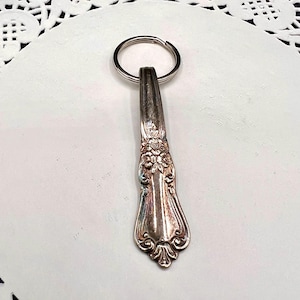 May include: Silverware keychain featuring a decorative floral design. The keychain is made from a repurposed spoon handle, with a silver metal key ring attached. The spoon handle has an ornate, vintage-style pattern.