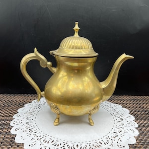 May include: A vintage brass teapot with a domed lid and a curved spout. The teapot has a decorative handle and sits on three small feet. The brass has a warm, golden color, and the teapot is placed on a white paper doily.