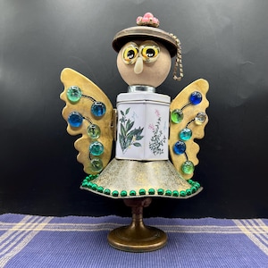 May include: A whimsical, handcrafted angel sculpture. It features a round head with owl-like eyes, a hat, and wings adorned with colorful glass beads. The body is made from a repurposed tea tin and a brass base.