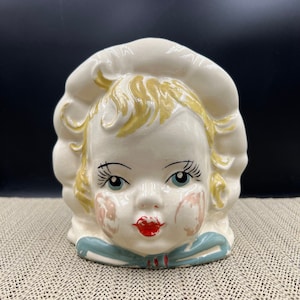 May include: Vintage ceramic head-shaped planter. The planter is off-white with a bonnet, blonde hair, blue eyes, and red lips. It has a blue bow at the neck and pink blush on the cheeks. A charming decorative piece.