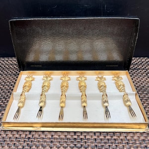 May include: A set of six gold-colored pineapple-shaped forks in a black box with gold trim. The forks are arranged neatly on a white surface inside the box. The box is open, revealing the forks and the text "FINE COLLECTION" and "24 K GOLD PLATED".