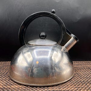 May include: A polished stainless steel stovetop kettle with a rounded dome shape. It features a black arched handle, a matching black knob on the removable lid, and a short, angled spout. The metallic surface reflects light, showing a smooth finish.