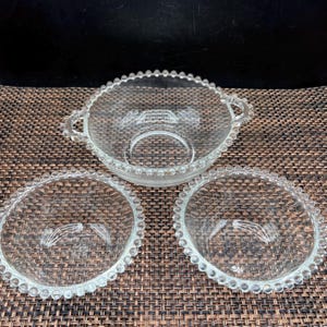 May include: Three clear glass bowls with a beaded edge. One bowl has two handles. The bowls are arranged on a brown woven surface. The bowls are made of clear glass.