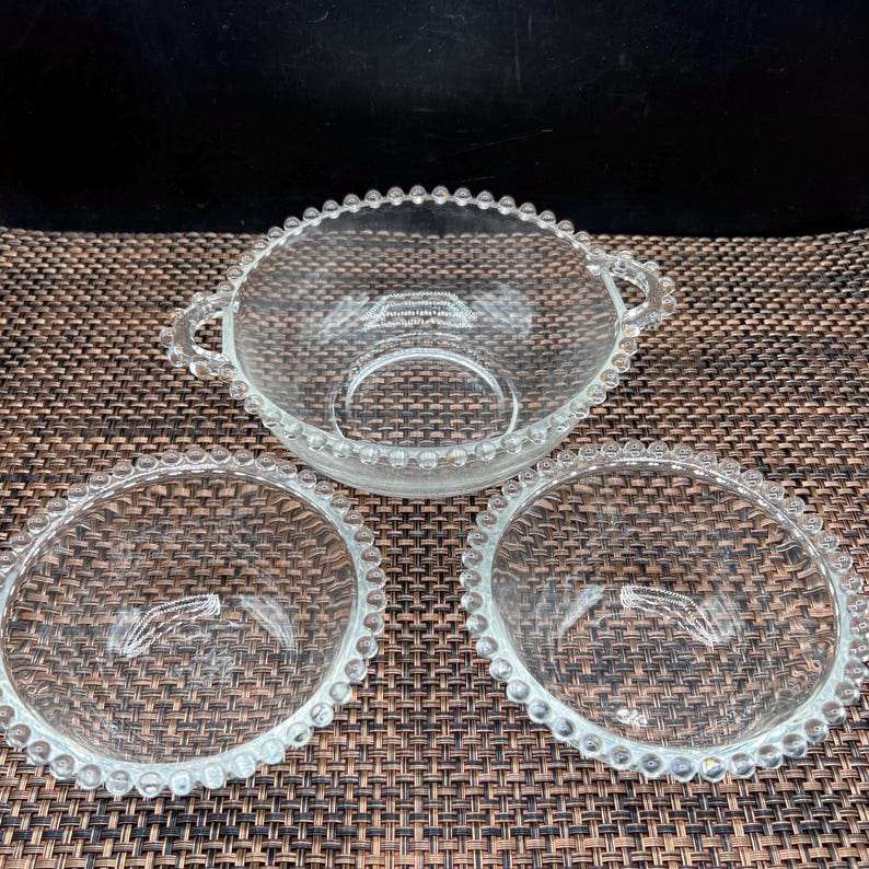 May include: Three clear glass bowls with a beaded edge. One bowl has two handles. The bowls are arranged on a brown woven surface. The bowls are made of clear glass.