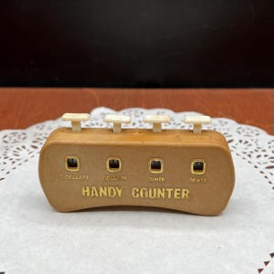 May include: A vintage brown plastic Handy Counter with four white buttons and four small square windows. The text "HANDY COUNTER" is printed in gold lettering. The windows are labeled "DOLLARS", "DIMES", and "CENTS".