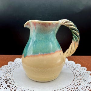 May include: A handmade ceramic pitcher with a twisted handle. The pitcher features a color gradient, transitioning from a tan base to a turquoise middle and a reddish-brown rim. The handle is a twisted rope design with green and tan hues.