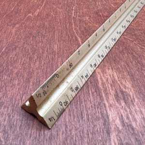 May include: A vintage ivory-colored triangular architect's scale ruler. The ruler features multiple scales with inch markings and numbers. The scale is made of a light-colored material and is set against a dark wood grain surface.