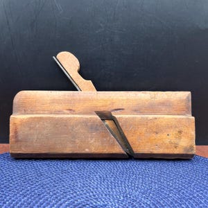 May include: A vintage wooden hand plane featuring a rectangular body with a natural wood grain finish. The tool includes a metal blade set at an angle within a central slot, secured by a wooden wedge component extending upward from the main body.
