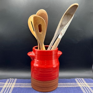 May include: A red and brown ceramic utensil holder with wooden spoons and a knife inside. The holder has two small handles and a two-tone design. The wooden utensils include a slotted spoon and a spatula.