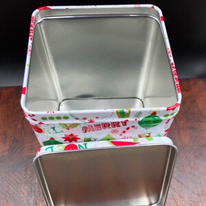 May include: Open rectangular Christmas tin with a lid. The tin is white with a festive pattern of ornaments, candy canes, and the word "MERRY" in red and green. The interior is silver.