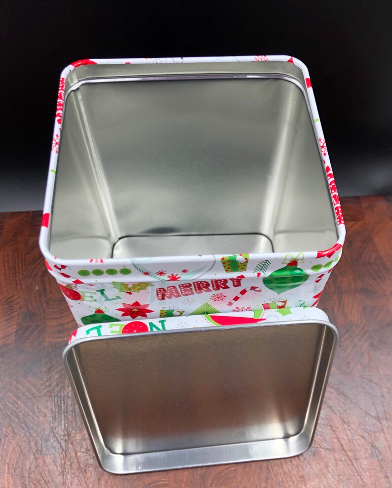 May include: Open rectangular Christmas tin with a lid. The tin is white with a festive pattern of ornaments, candy canes, and the word "MERRY" in red and green. The interior is silver.