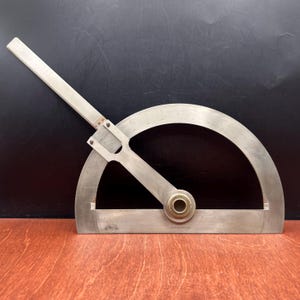 May include: A silver-toned metal protractor with a semi-circular base and a pivoting arm. The tool features engraved degree markings along the curved edge and a central brass-colored circular pivot point. The arm extends beyond the base.