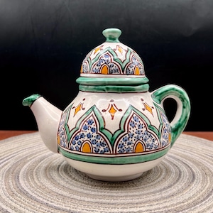 May include: A ceramic teapot with a removable lid, featuring a white base decorated with hand-painted green, blue, and yellow geometric patterns. The teapot has a curved handle, a short spout, and a rounded body with a matching domed lid.