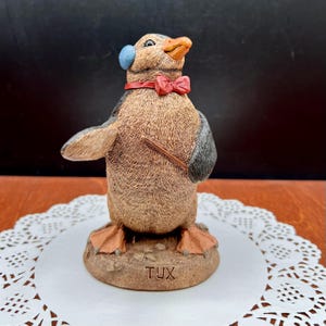 May include: A whimsical penguin figurine, dressed in a tuxedo, with a red bow tie and a blue ear covering. The penguin holds a baton and is standing on a base that reads "TUX". The figurine has a detailed, textured appearance.