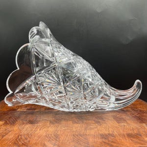 May include: A clear glass cornucopia vase featuring a prominent diamond-cut pattern across its curved body. The wide, scalloped opening transitions into a tapered, pointed end, with intricate geometric facets reflecting light throughout the piece.