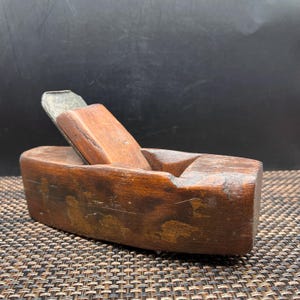 May include: A vintage hand plane constructed from dark brown wood with a weathered finish. The tool features a rectangular wooden body and a metallic blade secured by a wooden wedge, showing signs of age and use on the surface of the wood.