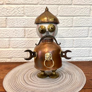 May include: A decorative robot figurine constructed from mixed metal components. The head is a baseball with a brass helmet, brass eyes, and a metal mustache. The body is a copper vessel featuring a gold lion head door knocker and wrench arms.