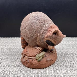 May include: A brown and gray raccoon figurine with a black eye mask, positioned on a textured base with a small green frog. The base features the text '92' and 'CLOSE ENCOUNTER' embossed on the front, depicting a scene of wildlife interaction.