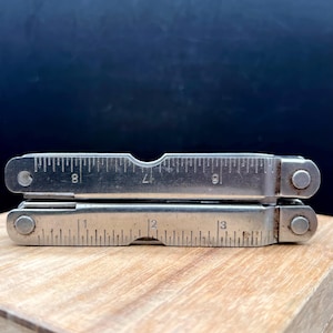 May include: A silver-colored metal multi-tool in a folded position. The tool features integrated ruler markings along the edges, with the bottom section showing measurements from 1 to 3 inches and the top section showing 6 to 8 inches.