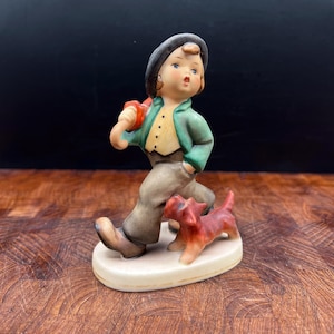 May include: A porcelain figurine of a young boy walking with a red dog. The boy is wearing a green jacket, tan pants, and a black hat. The dog is red and orange. The figurine is on a white oval base.
