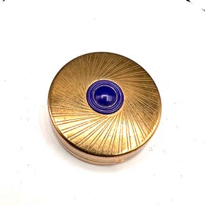 May include: A round, gold-toned compact with a sunburst design radiating from a central, deep blue, circular embellishment. The compact has a hinged lid and a smooth, polished finish, suggesting a vintage or antique style.