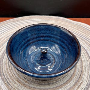 May include: A handmade, blue ceramic shaving bowl with a central post for holding a shaving soap puck. The bowl has a dark brown rim and a textured, glazed interior. The bowl is sitting on a neutral-toned, woven placemat.