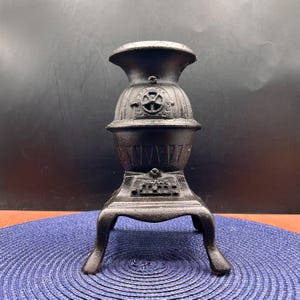 May include: A small, black cast iron stove with a decorative design. The stove has a rounded body, a flared top, and three curved legs. The front of the stove features a series of vertical lines and the word "FAVRE" is visible.