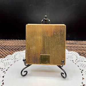 May include: A square gold-toned metal compact case featuring a vertical ribbed texture across the front surface and a small smooth rectangular panel at the bottom center, displayed on a black metal stand with curved legs and a top hook.