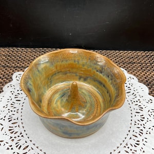 May include: A handmade ceramic apple baker with a central spike. The bowl has a wavy rim and is glazed in shades of brown, blue, and green. The baker sits on a white paper doily.