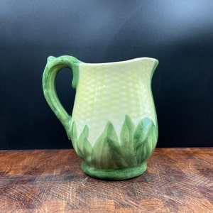 May include: A ceramic pitcher with a light yellow body and a green handle. The pitcher is decorated with green leaf designs around the base and a textured pattern on the body. The handle has a decorative, leaf-like detail.