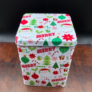 May include: A white rectangular Christmas tin with a festive pattern of red and green ornaments, Christmas trees, Santa faces, and the words "NOEL" and "MERRY". The tin is on a wooden surface.