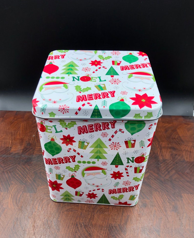 May include: A white rectangular Christmas tin with a festive pattern of red and green ornaments, Christmas trees, Santa faces, and the words "NOEL" and "MERRY". The tin is on a wooden surface.