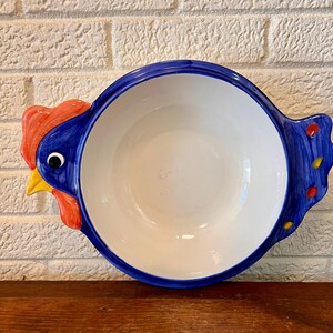 May include: A ceramic serving bowl with a rooster design. The bowl is white inside with a blue rim and blue handles shaped like a rooster's head and tail. The rooster's head is red and yellow, with a black eye.