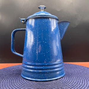 May include: A blue enamel coffee pot with white speckles. The pot has a handle, a spout, and a lid with a small knob. The pot is sitting on a blue placemat. A classic camping or kitchen item.