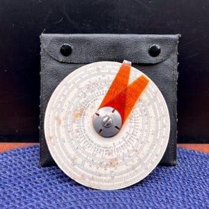 May include: A vintage circular slide rule with a white face, black markings, and an orange cursor. The slide rule is housed in a black leather case with two snap closures. The face has various scales and numbers.