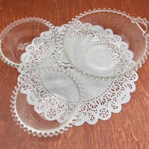 May include: Three clear glass serving bowls with beaded rims, resting on white lace doilies. The bowls have a simple, elegant design, perfect for serving appetizers or desserts. The background is a warm, wooden surface.