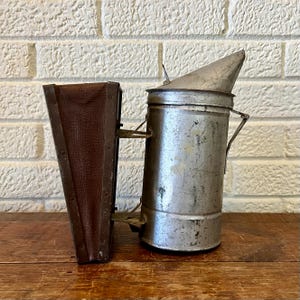 May include: An antique metal bee smoker with a conical top and a brown leather bellows. The smoker is cylindrical with a handle and a spout. The bellows is rectangular and folds in a zig-zag pattern. The items are on a wooden surface.