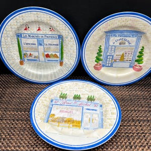 May include: Set of three decorative ceramic plates with blue rims. Each plate features a different French shop scene: a market, a bakery, and a patisserie. The plates have a brick-like background and are adorned with text in French.