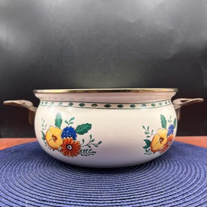 May include: A round, white enameled metal pot featuring a floral pattern with yellow, orange, and blue flowers and green leaves. The pot has a gold-toned metal rim and two metallic handles on the sides, with a green leaf border near the top edge.