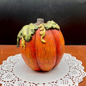 May include: A decorative pumpkin figurine featuring a vibrant orange body with a distinct black crackle finish. The top of the pumpkin is adorned with sculpted green leaves and a small stem, creating a textured appearance across the entire surface.