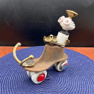 May include: A whimsical, handmade sculpture resembling a roller skate. It features a brown, shoe-shaped base with wheels made from white ceramic and red accents. The sculpture incorporates brass and gold-toned metal elements, along with a decorative head with pink-rimmed eyes.
