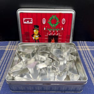 May include: A rectangular metal tin with a Christmas-themed lid featuring a fireplace scene and a cartoon peanut character. Inside the tin are several metal cookie cutters in various shapes, including stars and a reindeer. The tin is open, revealing the contents.