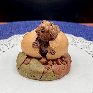 May include: A detailed figurine of a brown animal emerging from a cracked egg. The egg sits atop a base of green and brown leaves and small acorns. The figurine is likely a collectible or decorative item.