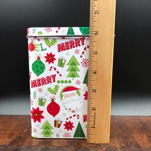 May include: A rectangular Christmas tin with a white background and a festive pattern of red, green, and white holiday elements. The tin features the words "MERRY" and "NOEL", along with images of Santa Claus, ornaments, and snowflakes. The tin is approximately 7 inches tall.
