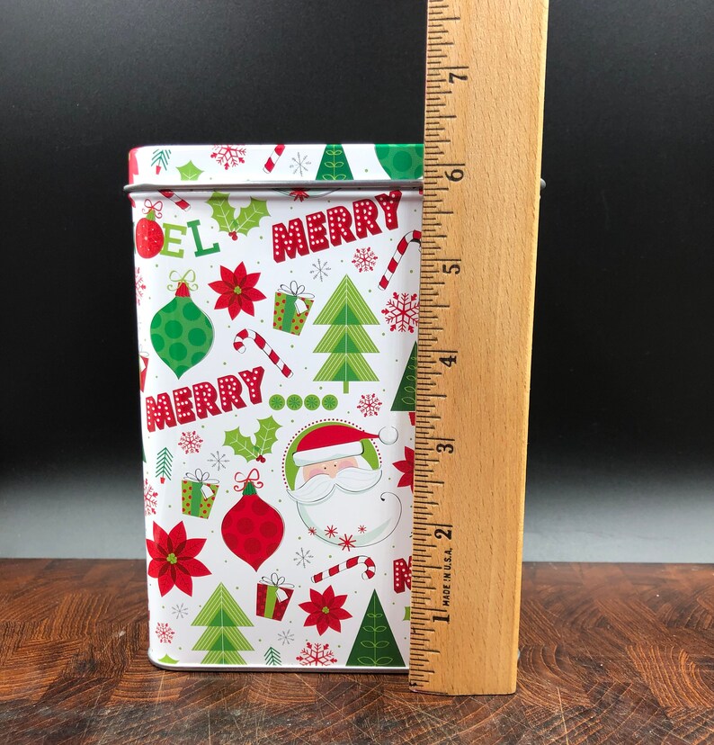 May include: A rectangular Christmas tin with a white background and a festive pattern of red, green, and white holiday elements. The tin features the words "MERRY" and "NOEL", along with images of Santa Claus, ornaments, and snowflakes. The tin is approximately 7 inches tall.