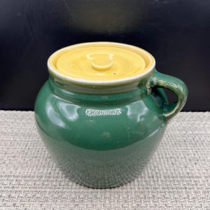 May include: A round, forest green ceramic crock with a single side handle and a smooth, glossy finish. The container features a circular, recessed yellow lid with a small central knob, creating a distinct contrast against the deep green body.