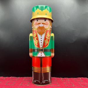 May include: A decorative, cylindrical nutcracker figurine. The figurine is painted with a green and red uniform, a brown beard, and a yellow crown. The design includes gold accents and a white shirt. The nutcracker is a festive holiday decoration.
