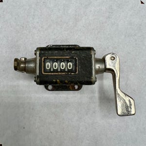 May include: An antique mechanical counter with a black body and a silver handle. The counter displays "0000" in white digits. The device has a metal base with two mounting holes and a silver lever on the side.